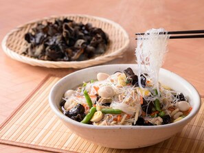 Un tazón de fideos con diversos vegetales y mariscos, acompañado de una cesta de mariscos secos al fondo.