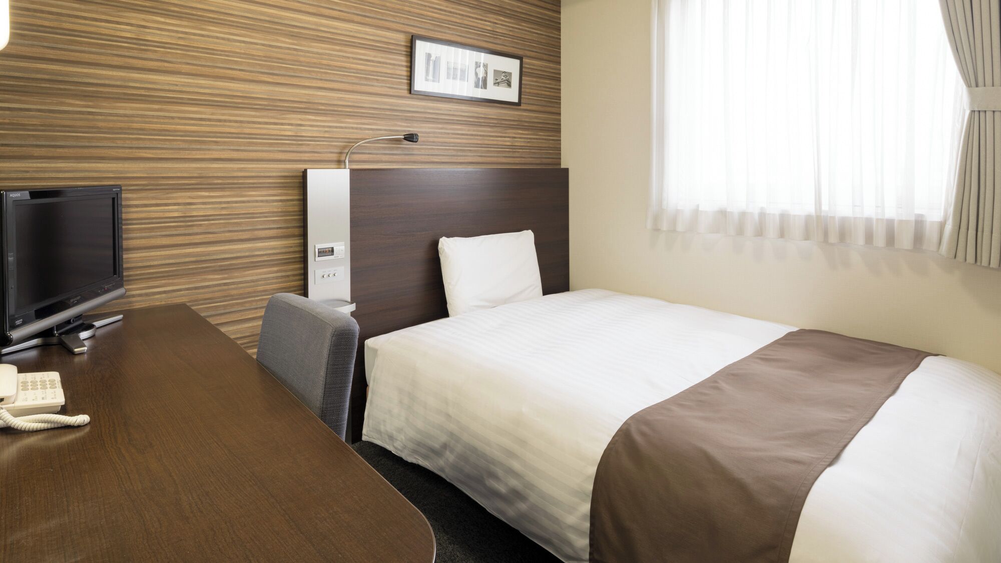 Una habitación de hotel con cama, escritorio con televisor, teléfono, lámpara y un cuadro en la pared.