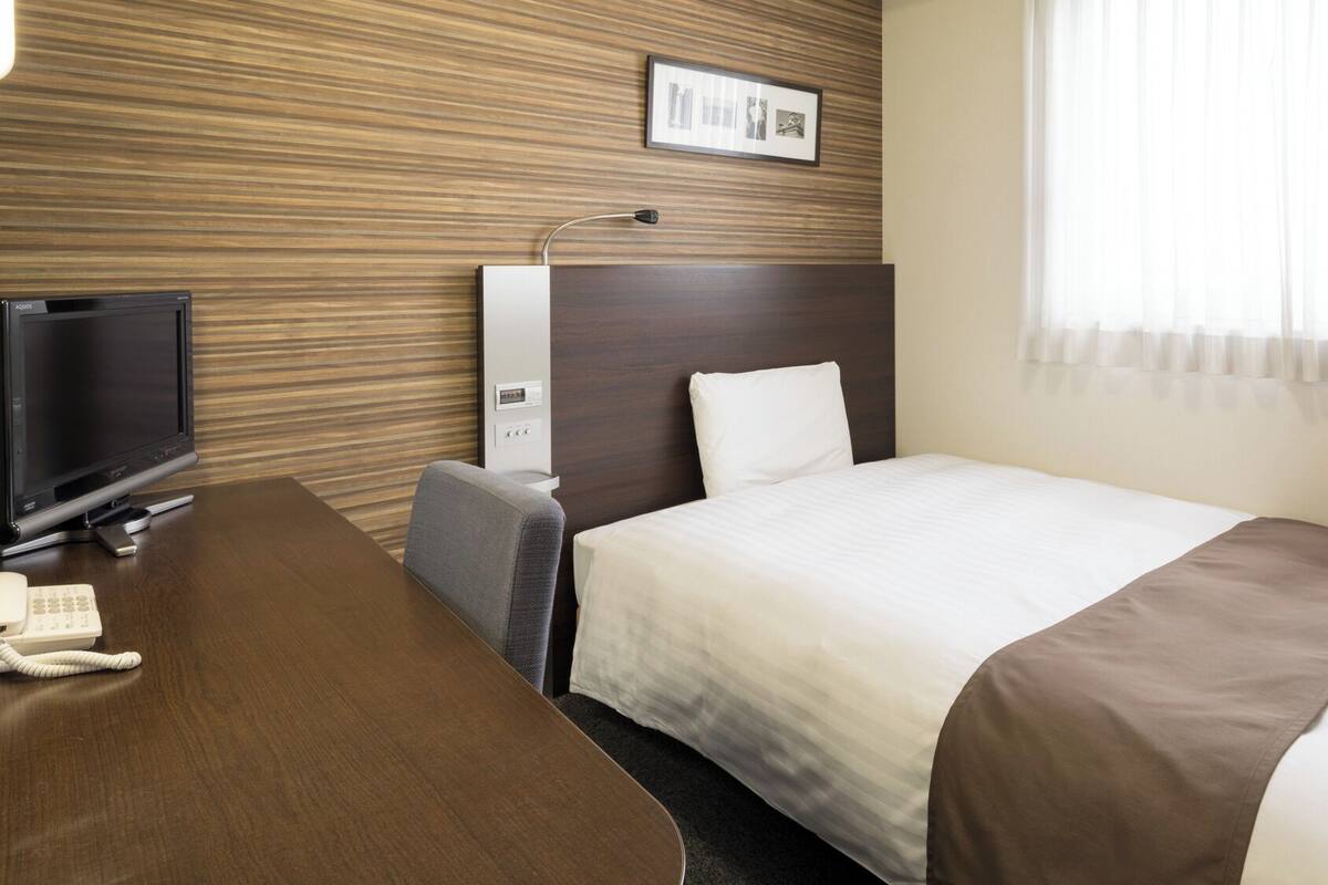 Una habitación de hotel con cama, escritorio con televisor, teléfono, lámpara y un cuadro en la pared.