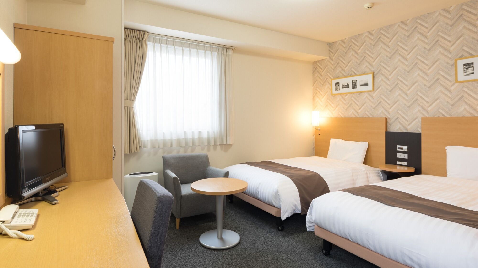 Habitación de hotel con dos camas, un escritorio con televisor y teléfono, una silla, una mesita y una ventana con cortinas.