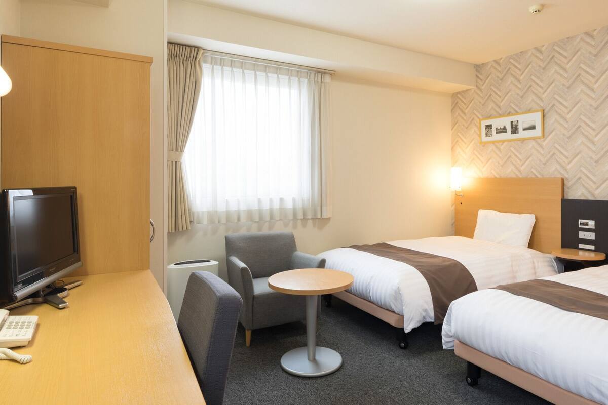 Habitación de hotel con dos camas, un escritorio con televisor y teléfono, una silla, una mesita y una ventana con cortinas.