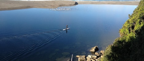 A clear view of a beach with calm waters, a person paddleboarding, and rocky terrain.