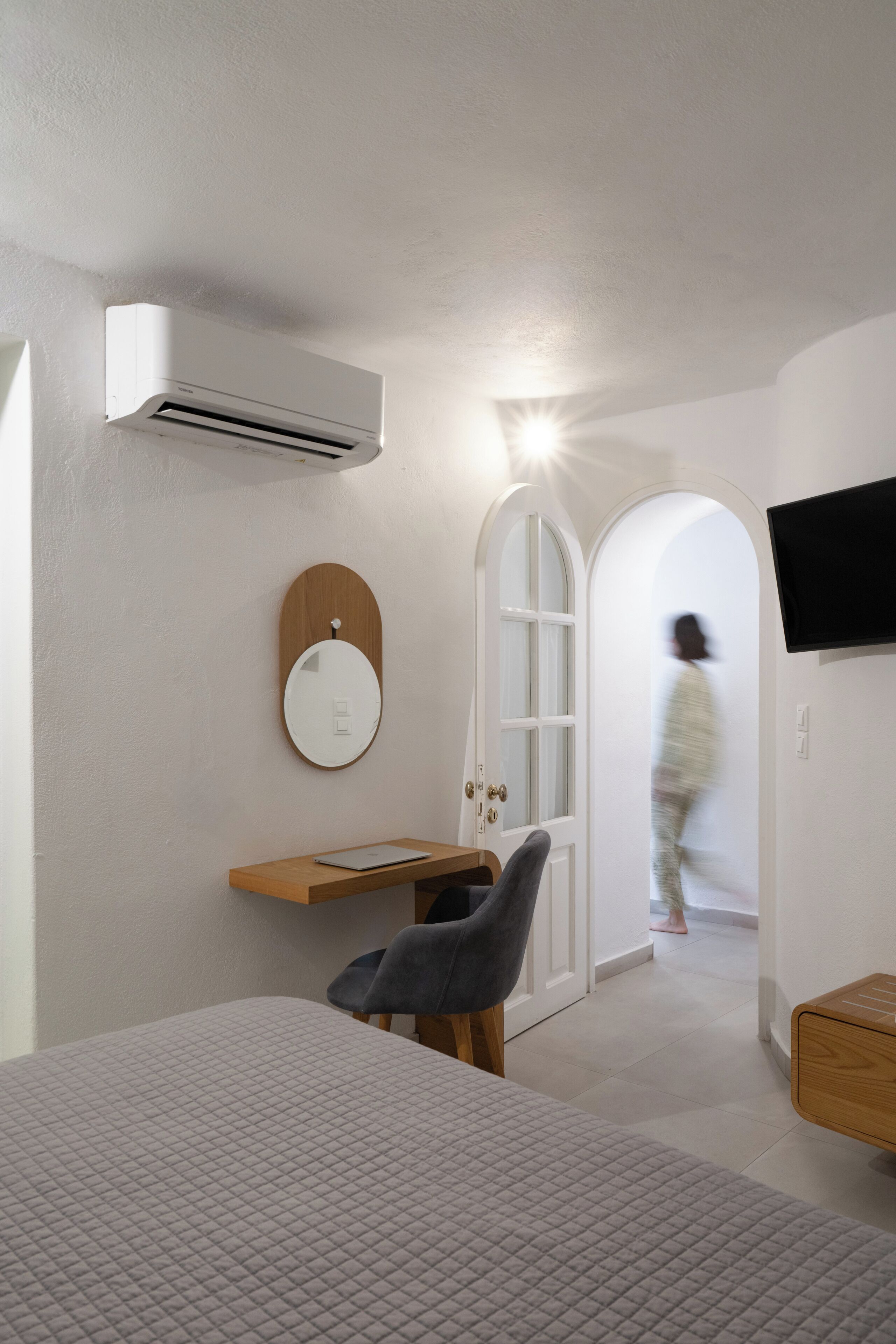 Une chambre à coucher comprenant un lit, un bureau, une chaise, un climatiseur, une télévision, un miroir et une porte donnant sur une autre pièce.