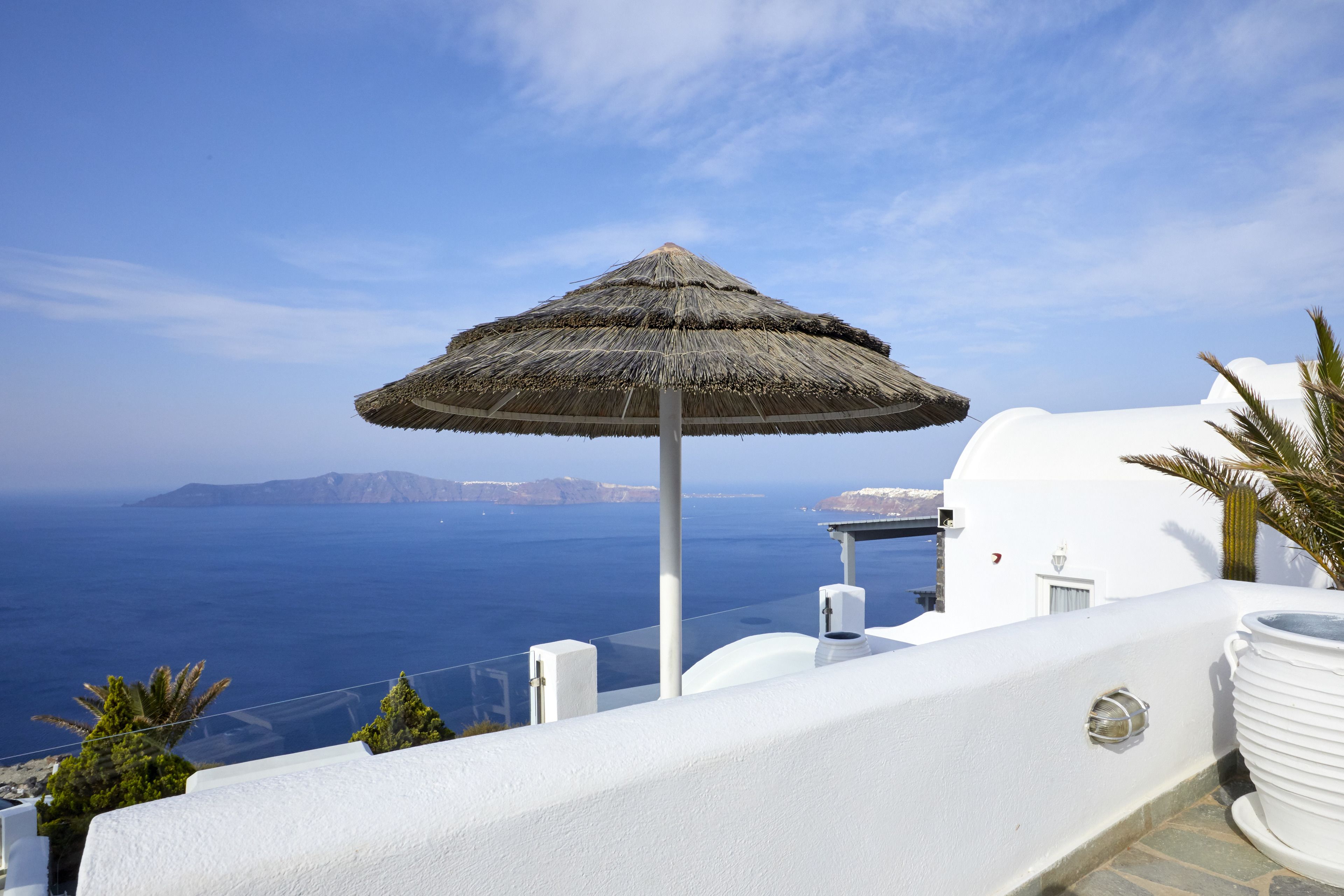 A white building with a thatched umbrella, overlooking a clear blue sea and distant land.