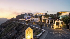 A coastal hotel with a stone pathway leading to a welcoming entrance, surrounded by Mediterranean-style buildings and a mountainous backdrop. - Santorini Princess Spa Hotel (Santorini)