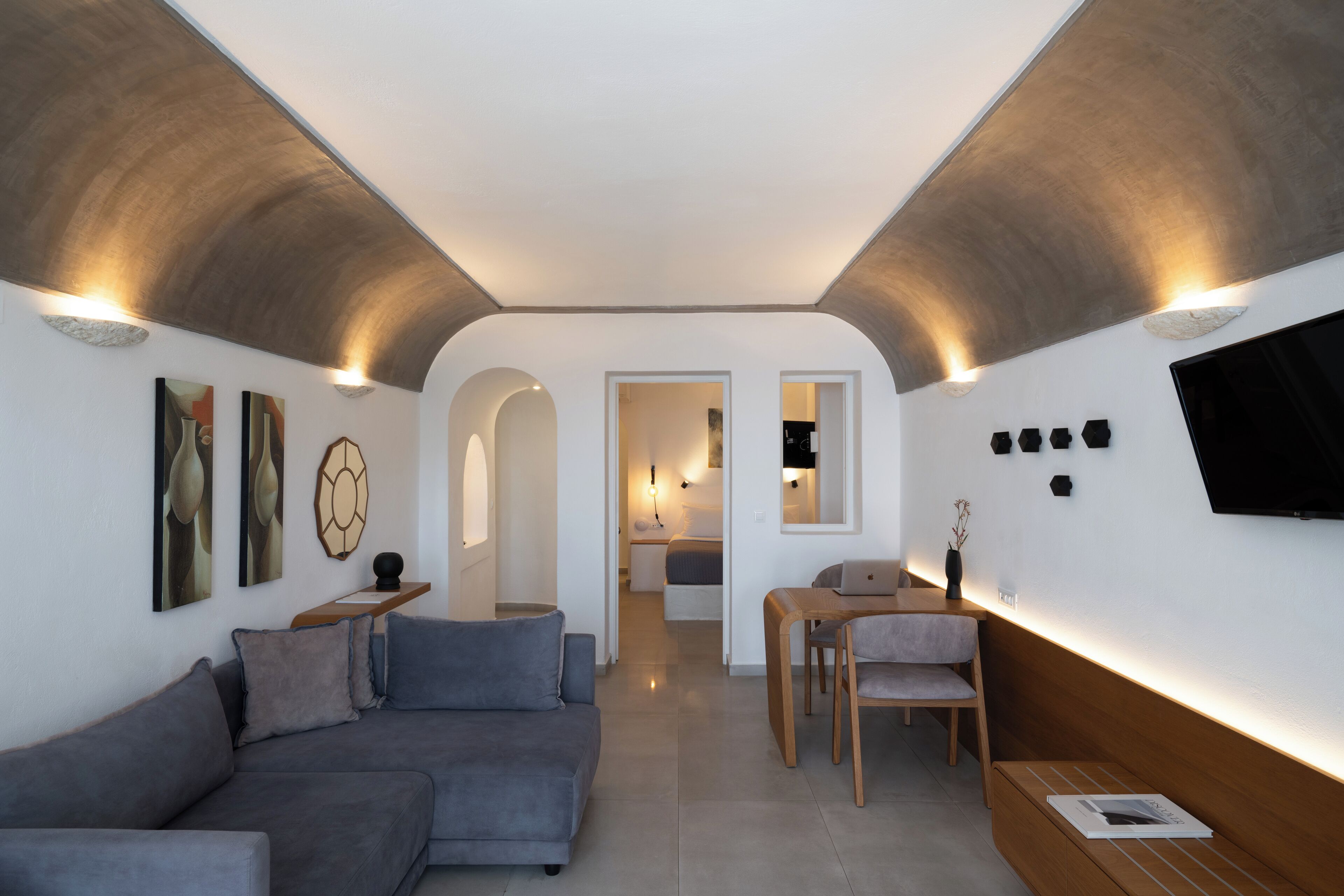 A modern living room with a grey sofa, a wooden dining table, and a TV mounted on the wall.