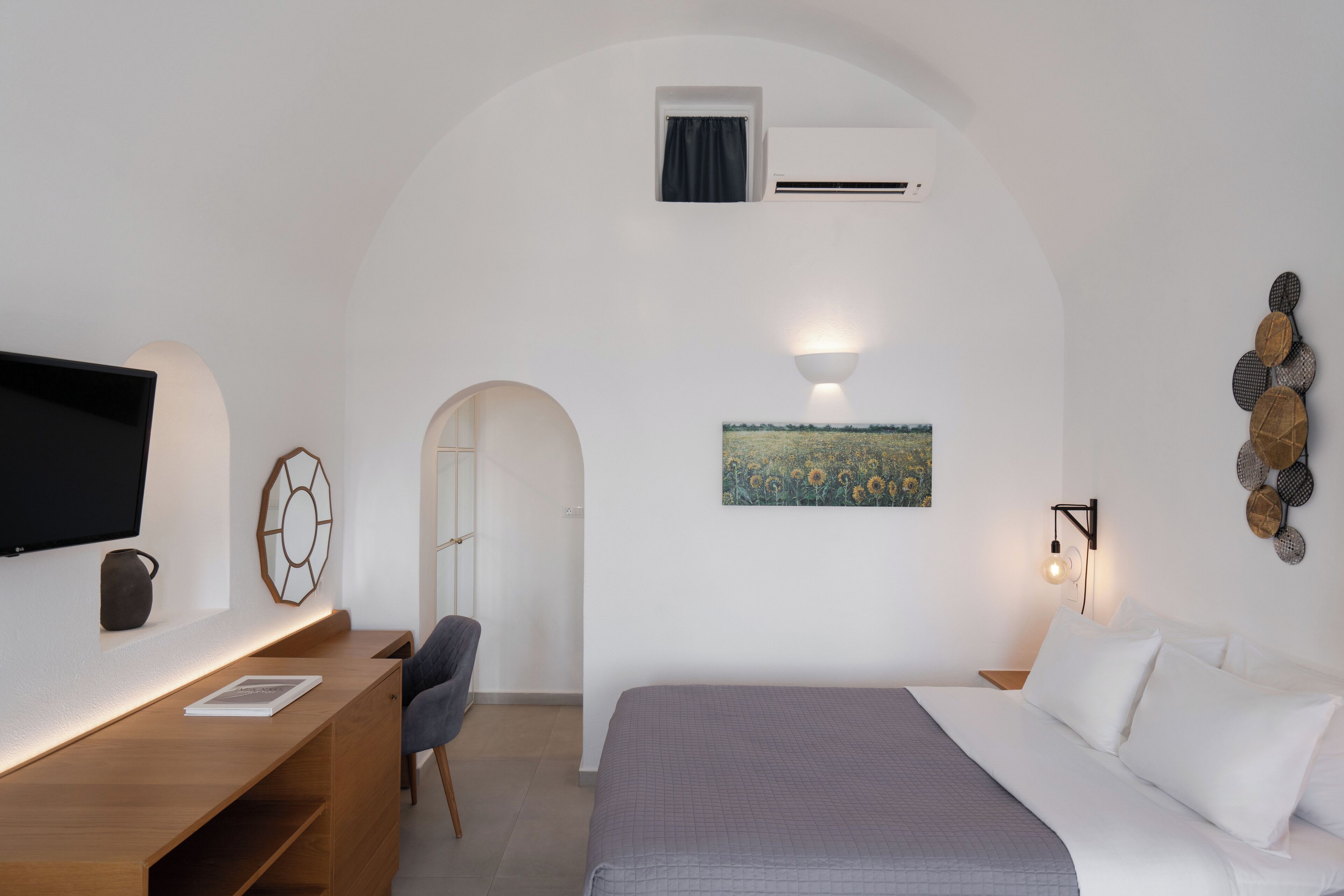 A modern hotel room with a flat-screen TV, a desk, a comfortable chair, a bed with white bedding, a painting of a field, and a wall-mounted light fixture.