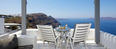 A covered outdoor seating area with white wicker furniture and a table, overlooking a coastal landscape with cliffs and the sea.