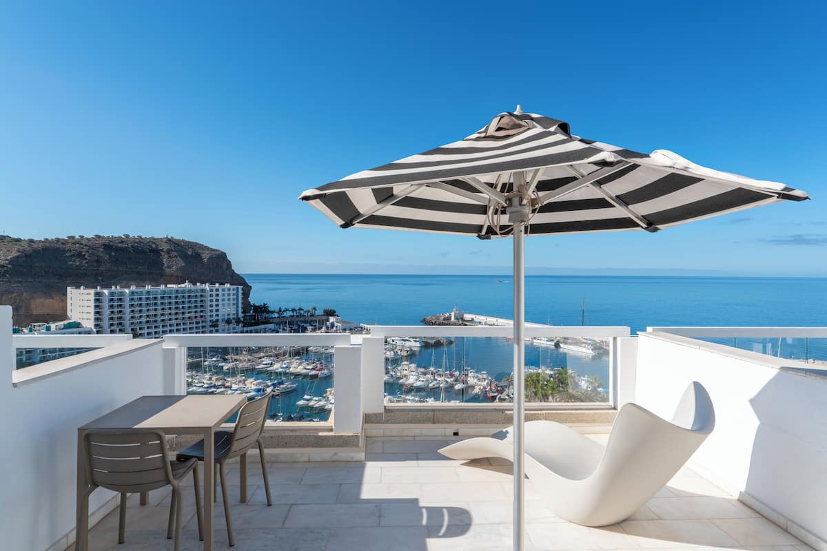 A balcony with a table, chairs, and a striped umbrella overlooking a marina and coastal buildings.