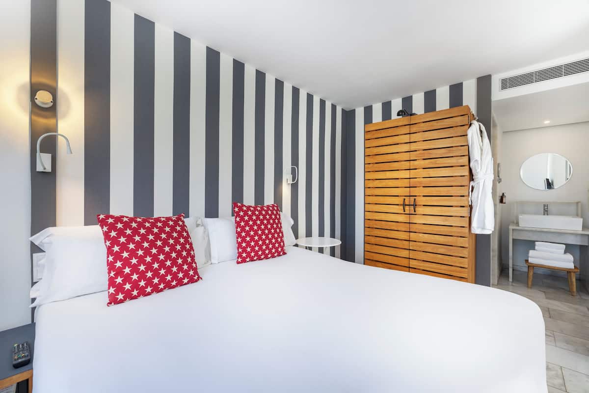 A hotel room with a large bed, striped walls, a wooden wardrobe, a small round table, and a bathroom with a mirror and sink.