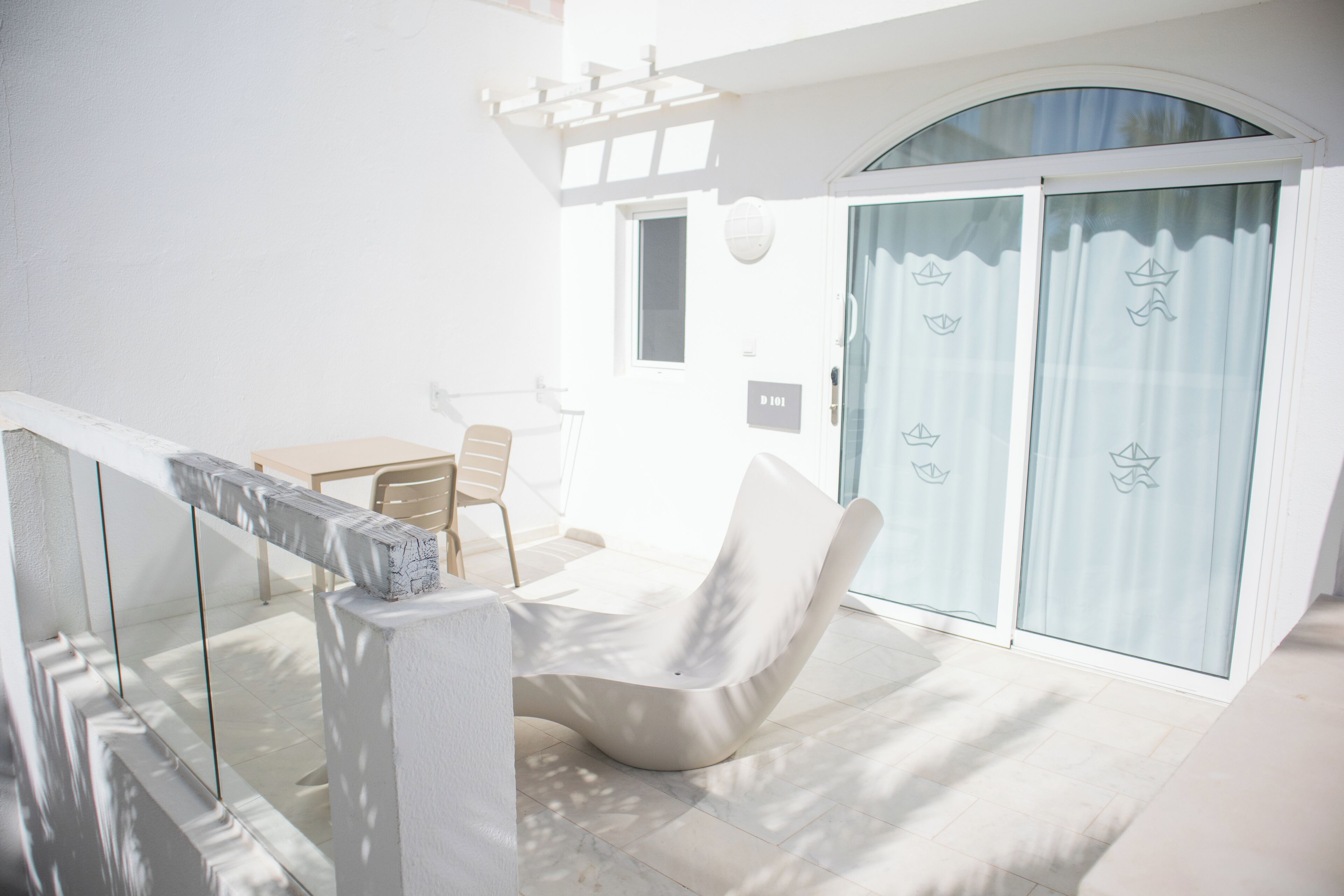 A bright, modern outdoor seating area with a white chair and table, a glass balustrade, and a sliding glass door with a nautical theme.