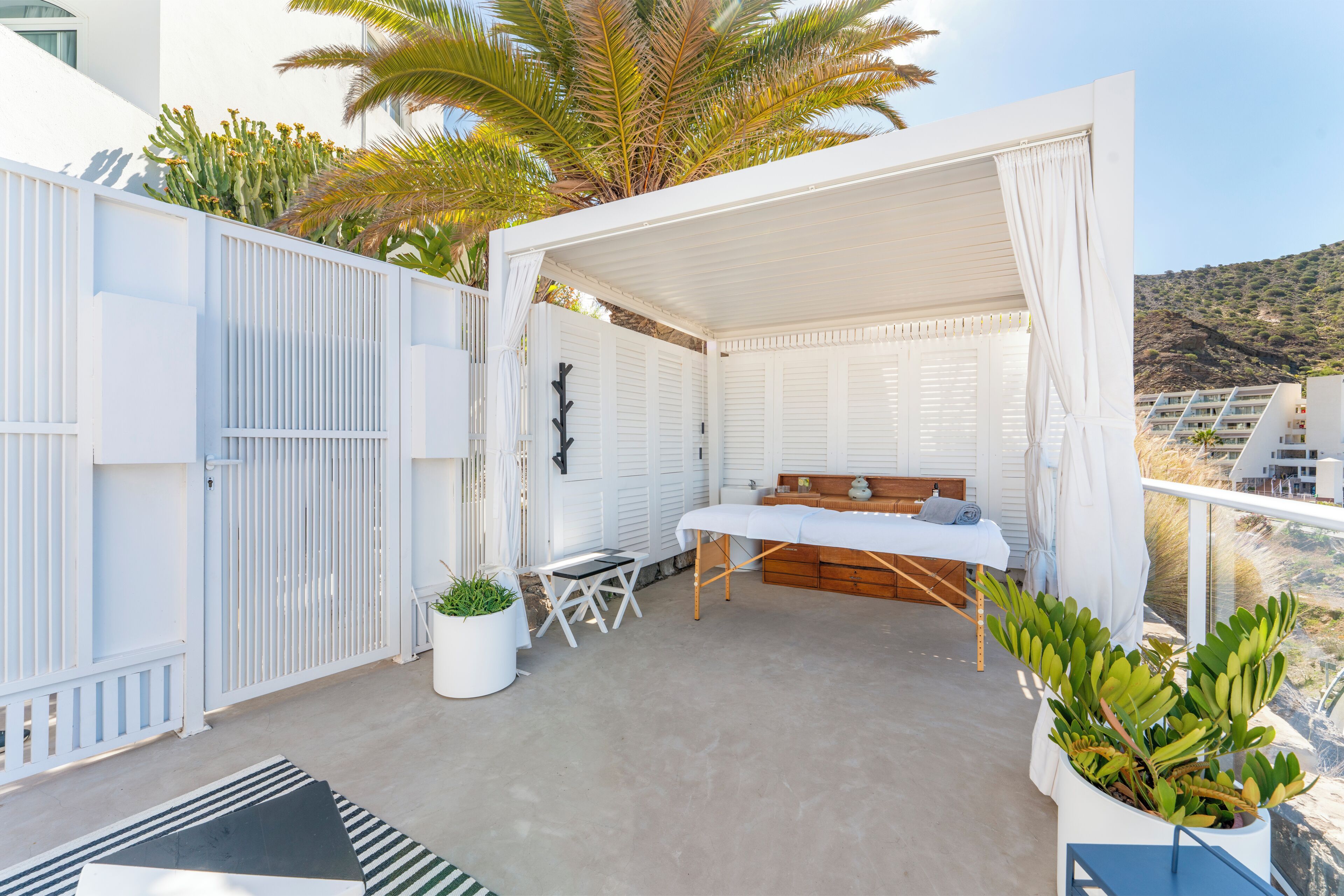A covered outdoor seating area with a bed, a table, and chairs, surrounded by white walls and plants.