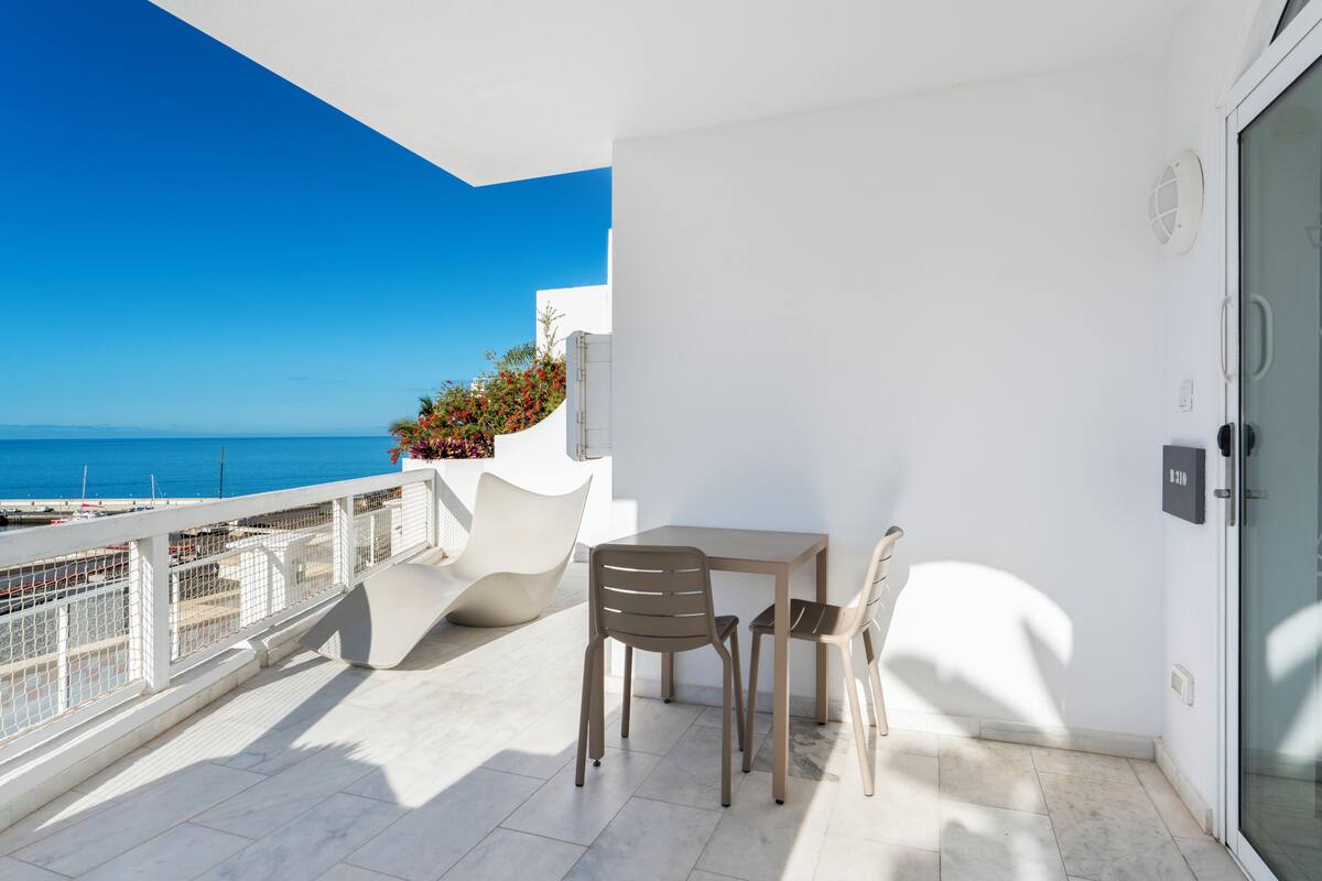 A balcony with a table and chairs, a view of the sea, and a white wall with a door.