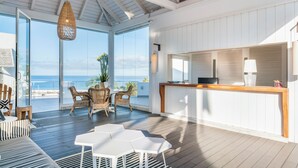 A spacious room with a wooden ceiling, large windows, a dining area with chairs, a white table, and a reception desk.