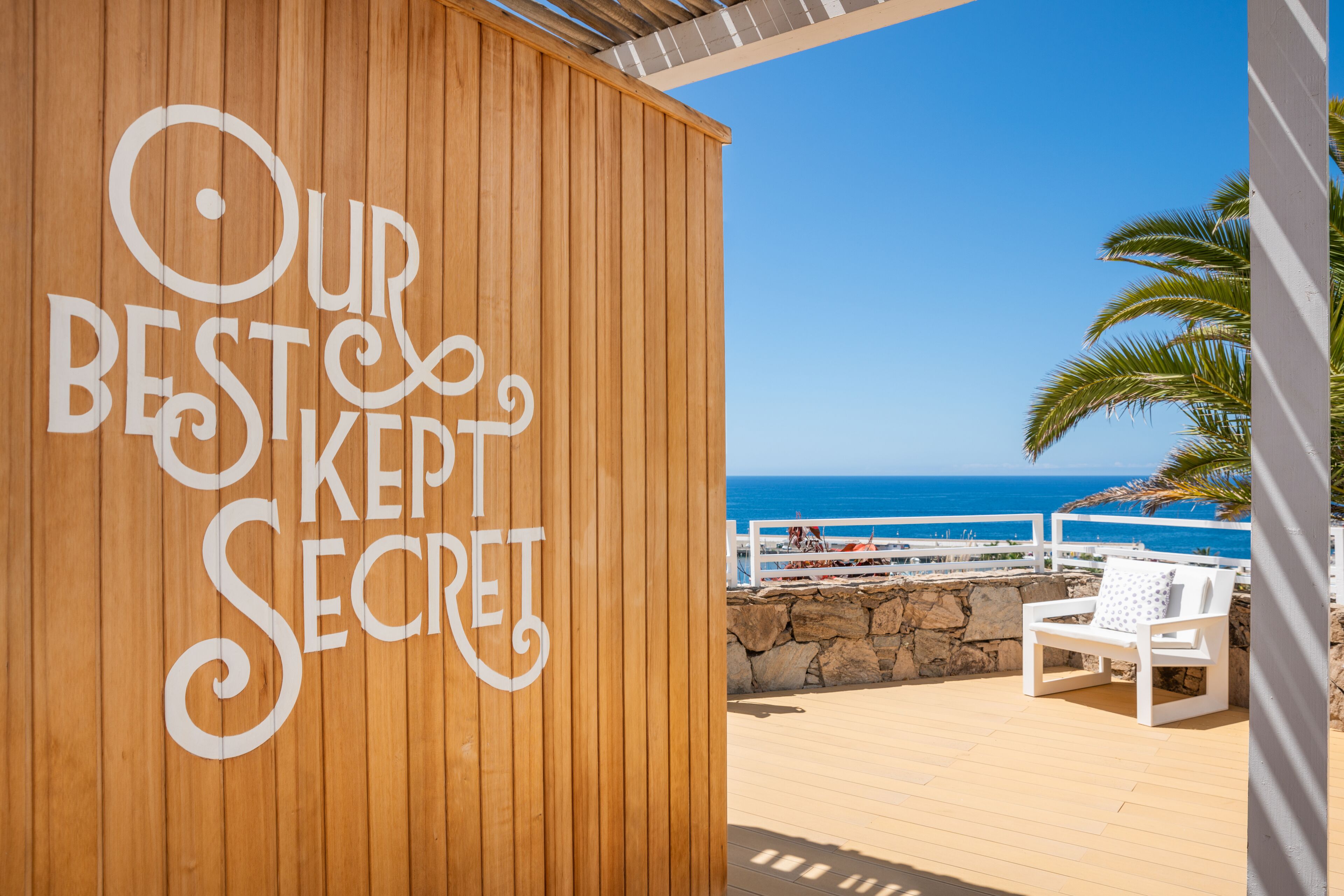 Un mur en bois sur lequel est inscrit le texte « NOTRE MEILLEUX SECRET » et une chaise blanche sur une terrasse en bois donnant sur la mer.