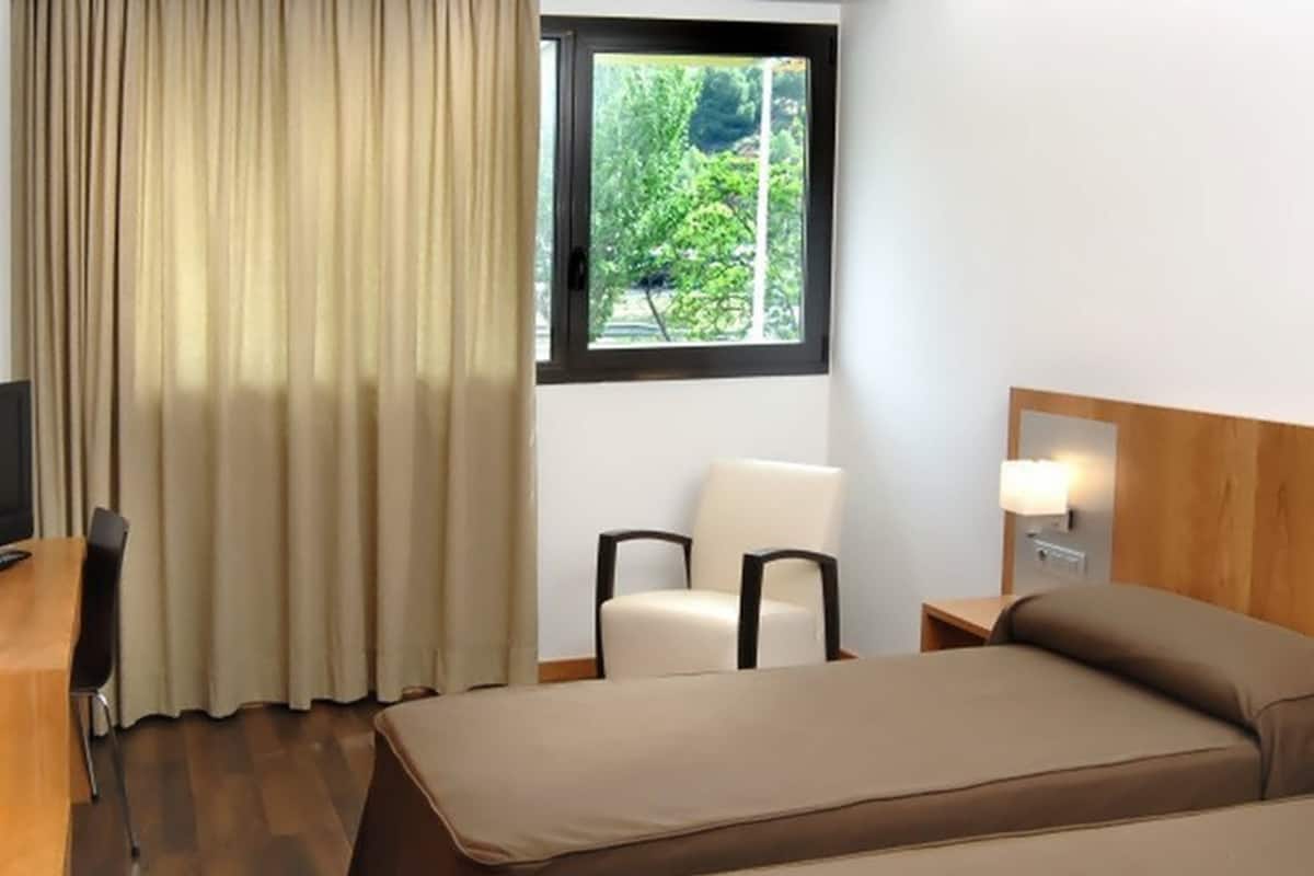 Une chambre d’hôtel avec deux lits, une chaise, un bureau, une lampe et une fenêtre donnant sur un paysage verdoyant.