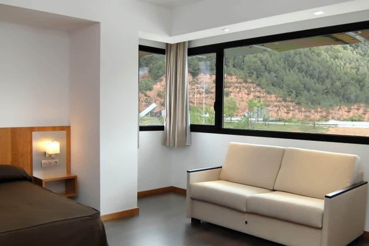 Une chambre d’hôtel moderne équipée d’un canapé, d’un lit, d’une table de chevet avec une lampe et d’une grande fenêtre donnant sur une zone boisée.