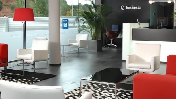 Un hall d’hôtel moderne avec un comptoir d’accueil, des chaises blanches, une lampe rouge et une grande fenêtre donnant sur un paysage verdoyant.