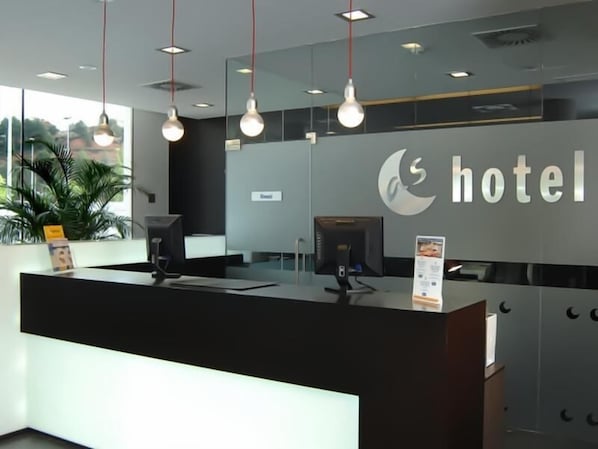 A hotel reception area with a modern design, featuring a reception desk, a digital screen, and a large window offering a view of the outdoors.