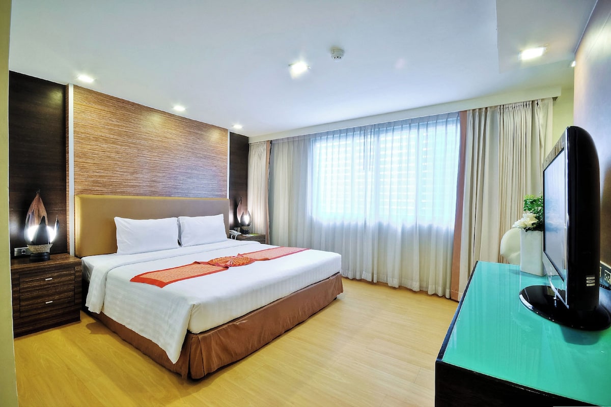 Habitación de hotel moderna con una cama grande, cabecera de madera, un televisor de pantalla plana y un ventanal con cortinas.