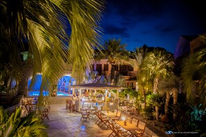 Un patio al aire libre con asientos y un bar, rodeado de palmeras y una piscina al fondo.