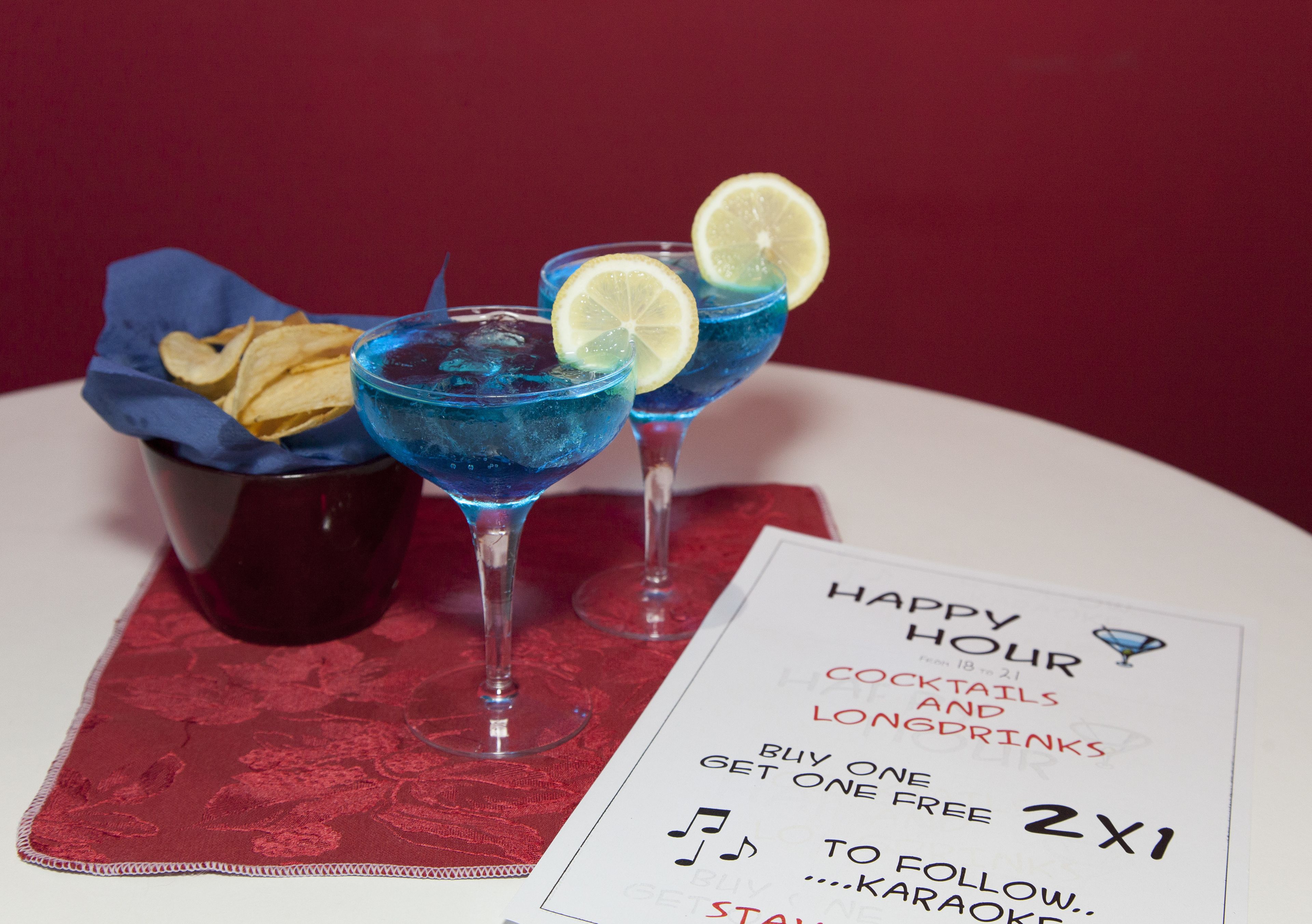 Three blue cocktails with lemon slices, a basket of chips, and a menu offering happy hour deals.