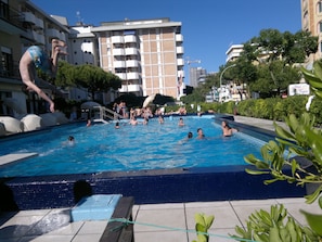 A swimming pool with people enjoying the water, surrounded by buildings and greenery. - Hotel Amalfi (Jesolo)
