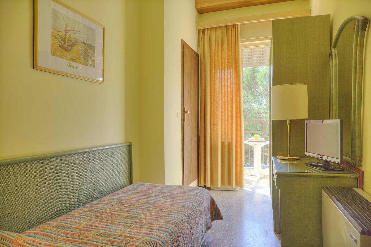 A hotel room with a bed, a desk with a computer, a window with curtains, and a framed picture on the wall.