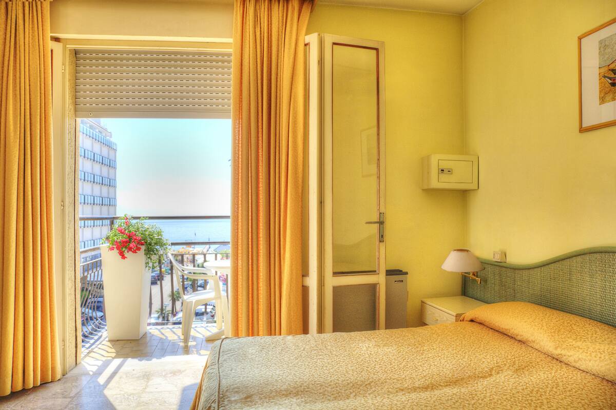 A hotel room with a bed, a nightstand, a lamp, and a view of the sea through a balcony door.