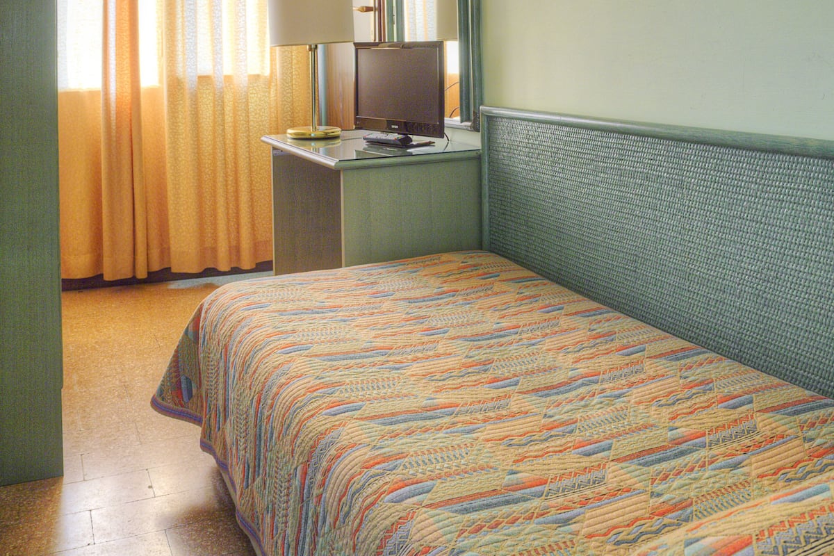 A hotel room with a bed, a desk with a computer, a lamp, a window with curtains, and a painting on the wall.
