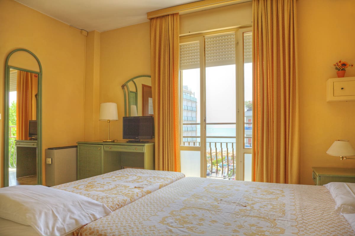 A hotel room with a bed, a television on a desk, a balcony with curtains, and a view of the sea.