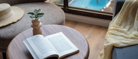 A room with a round wooden table, a book, a small plant, a straw hat, and a view of a swimming pool through a window.