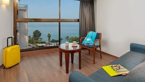 Habitación de hotel con un ventanal grande, suelo de madera, una maleta amarilla, una silla azul, una mesita redonda con bandeja y un sofá.