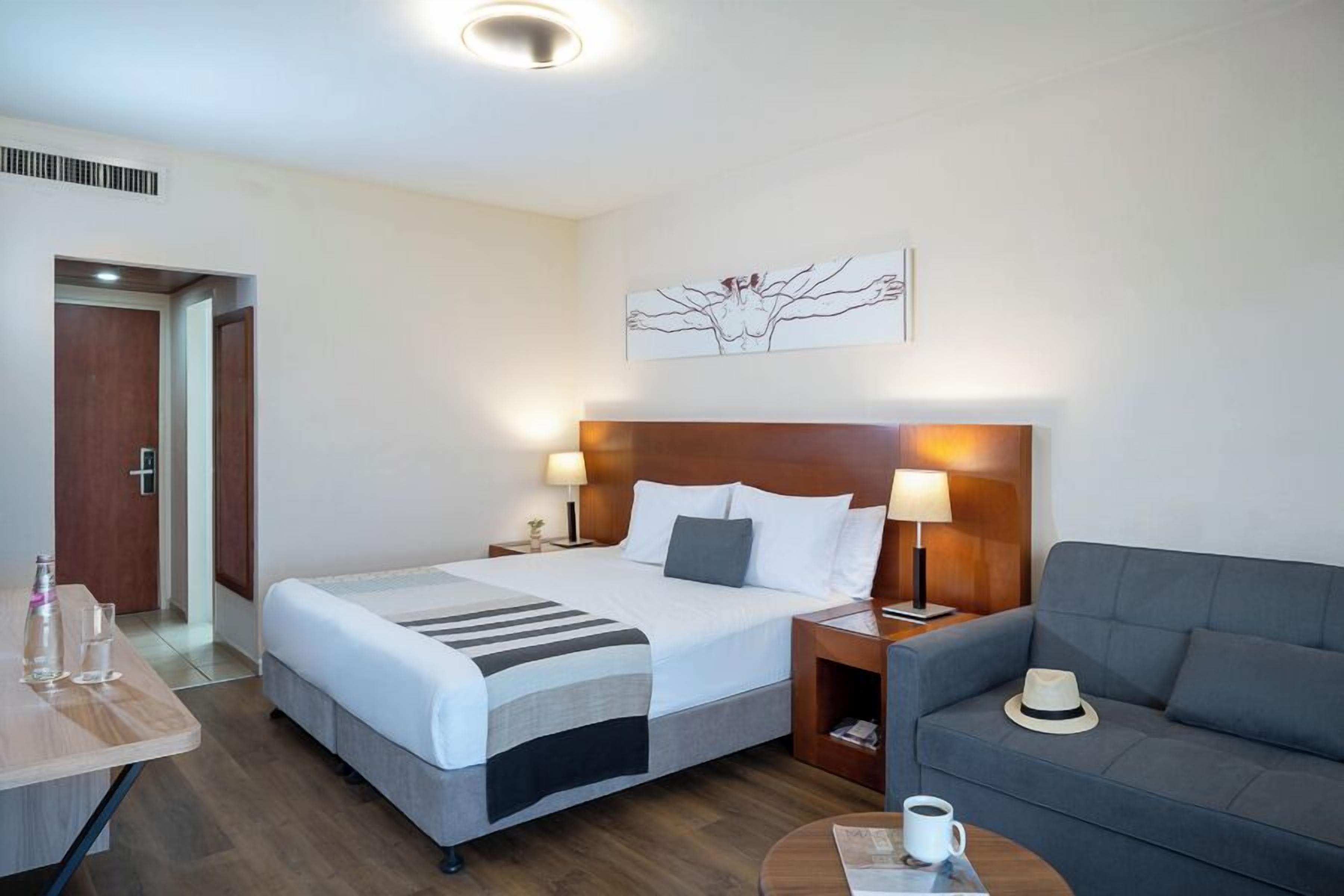 A hotel room with a bed, bedside tables, a sofa, and a painting on the wall.