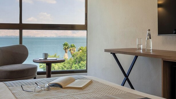 Habitación de hotel con un ventanal grande, una cama, una mesita con vasos y un libro, y un televisor en la pared.