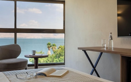 A hotel room with a large window, a bed, a small table with glasses and a book, and a TV on the wall.
