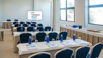 A conference room with tables set up for a meeting, each with a laptop, water bottle, and notepad. A screen displays the text "Cabogata Beach".