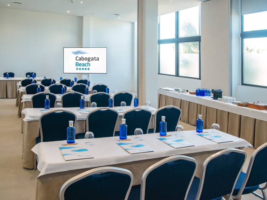 A conference room with tables set up for a meeting, each with a laptop, water bottle, and notepad. A screen displays the text "Cabogata Beach".