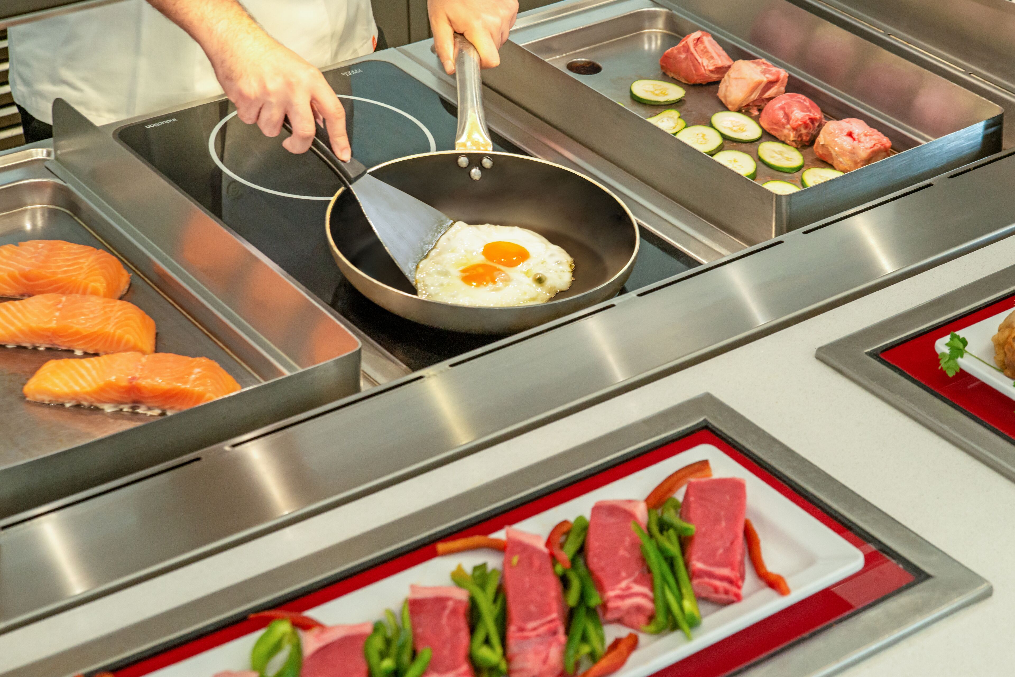 A kitchen with a griddle, a pan with eggs, and various meats and vegetables on display.