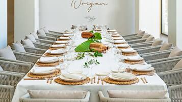 A long dining table set with white tablecloths, plates, glasses, and cutlery, surrounded by wicker chairs.