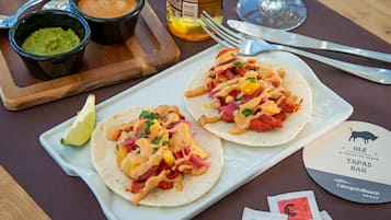 Dos tacos sobre un plato blanco, con una cuña de lima, acompañados por dos recipientes pequeños de salsa y guacamole sobre una tabla de madera.