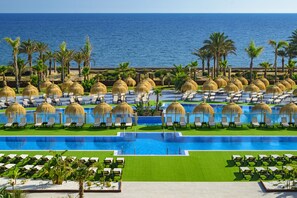 Zona de piscina en un resort con múltiples tumbonas y sombrillas de paja, rodeada de palmeras y vista al océano.