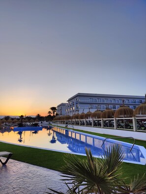 A hotel with a swimming pool, palm trees, and a sunset in the background.