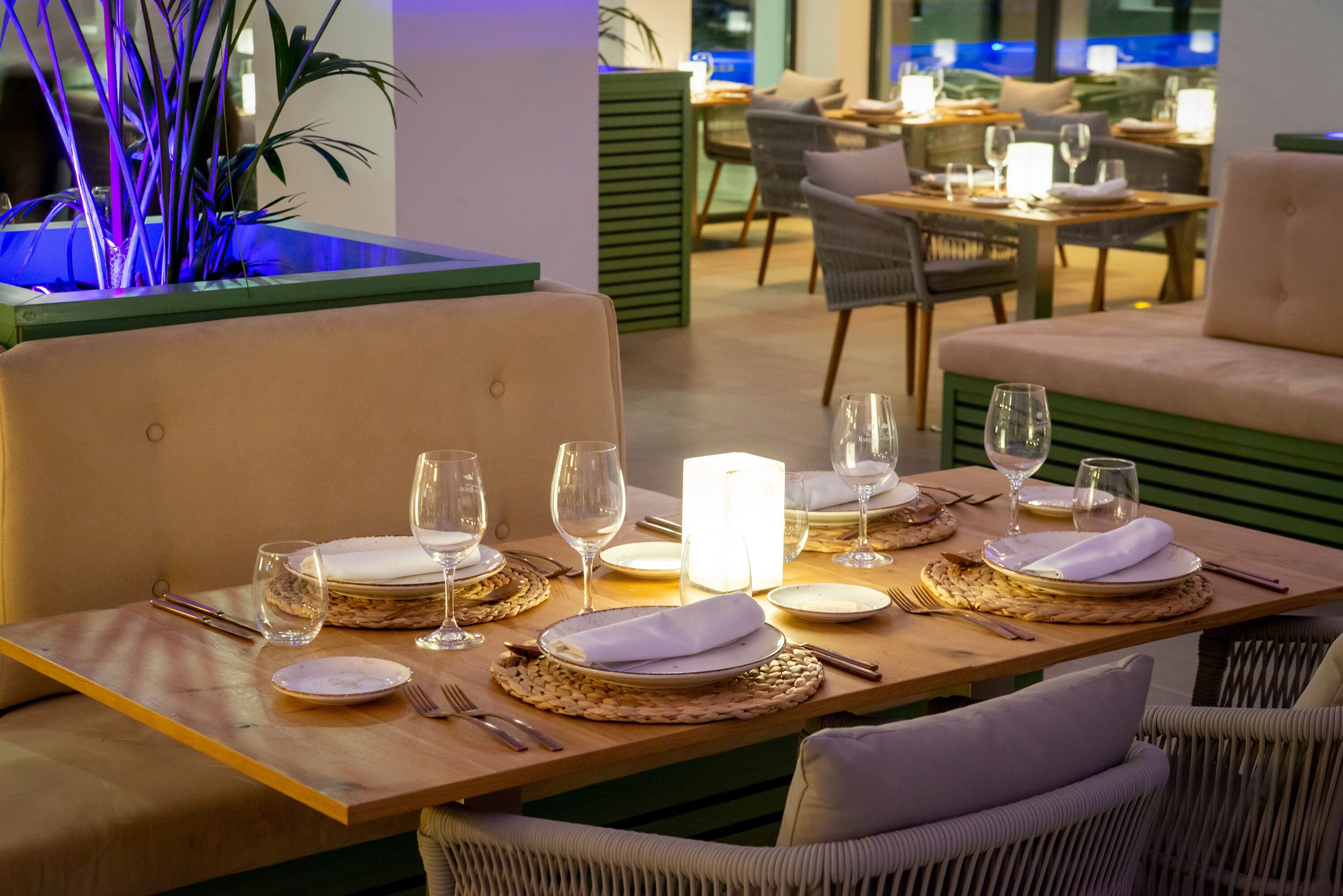 A dining area with tables set for two, featuring woven placemats, white napkins, and clear glassware.