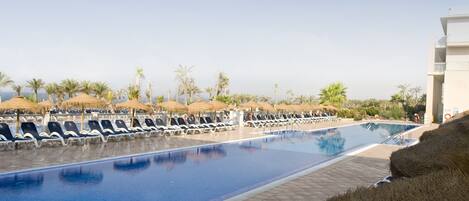 A pool area with lounge chairs and umbrellas, surrounded by palm trees and a clear sky.