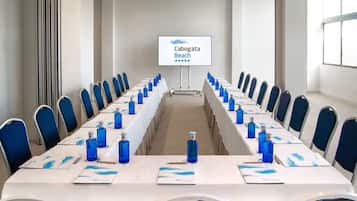 Un salón de conferencias con una larga mesa, sillas azules y una pantalla que muestra