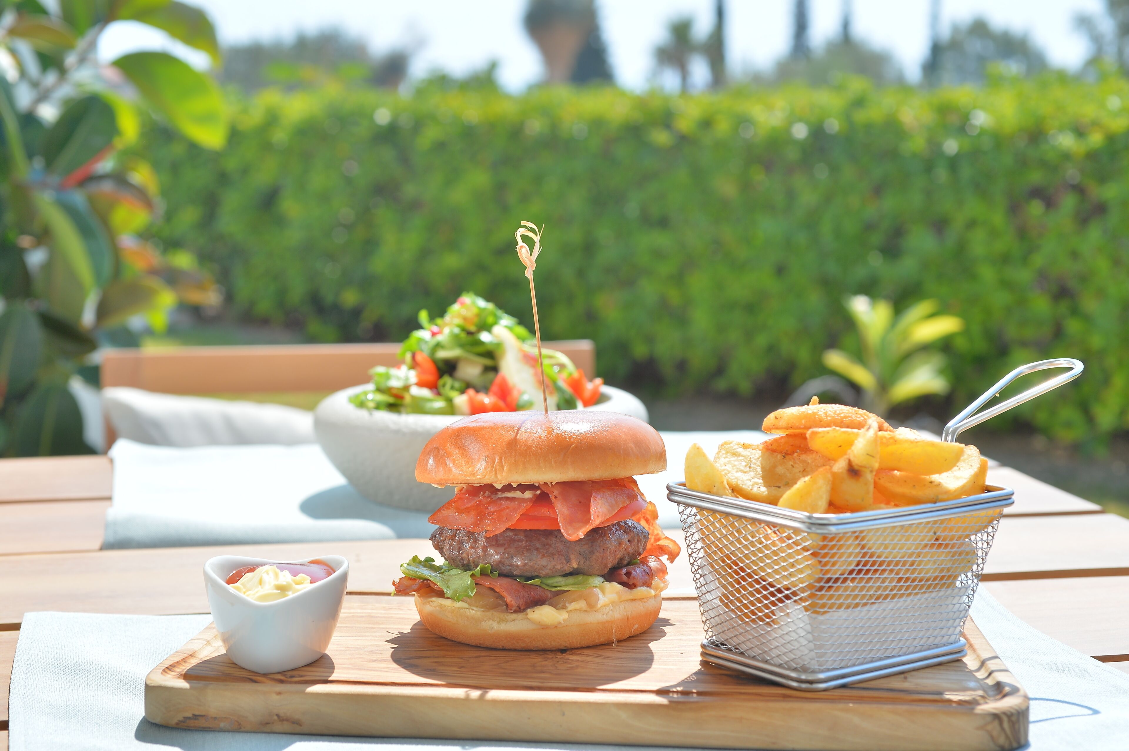 Ein Burger mit Speck, Salat und Tomate auf einem Brötchen, eine Salatschüssel und ein Korb Pommes auf einem Holztisch.