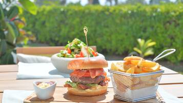 Ein Burger mit Speck, Salat und Tomate auf einem Brötchen, eine Salatschüssel und ein Korb Pommes auf einem Holztisch.