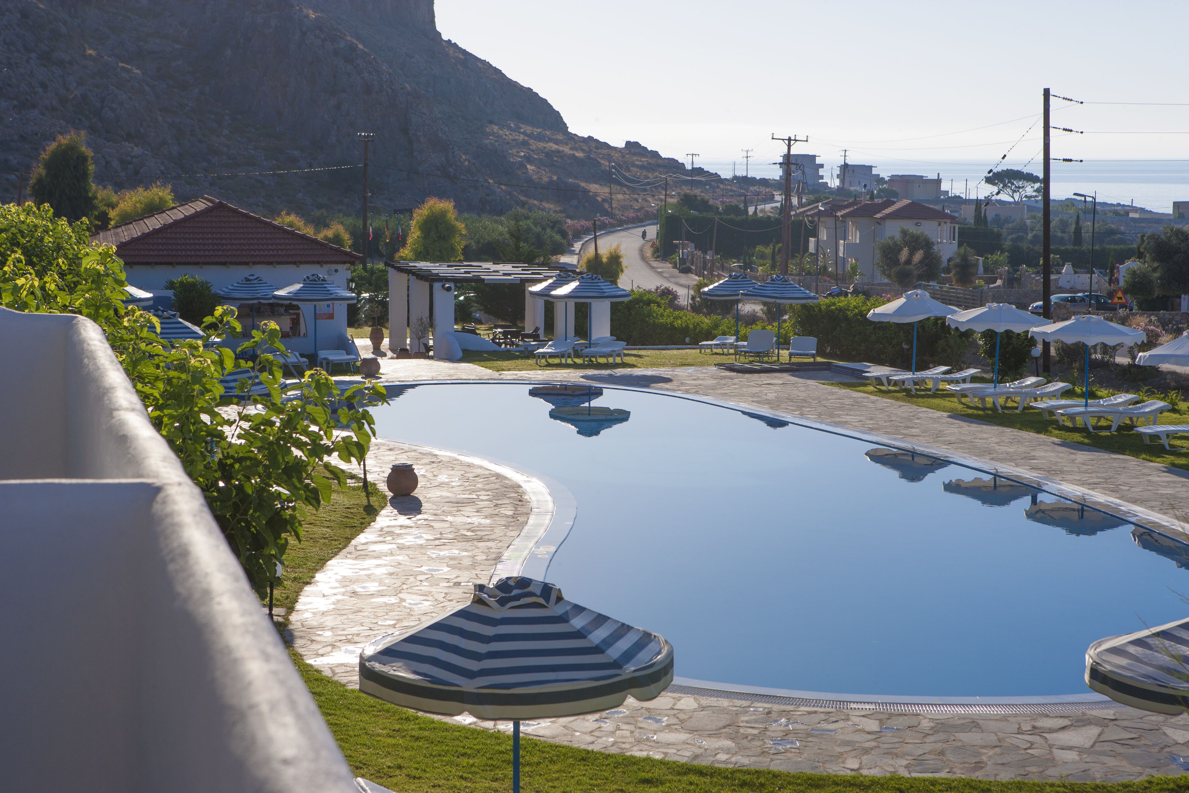 Piscina con sillones de descanso y sombrillas, rodeada de edificios y montañas al fondo.