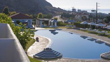 Piscina con sillones de descanso y sombrillas, rodeada de edificios y montañas al fondo.